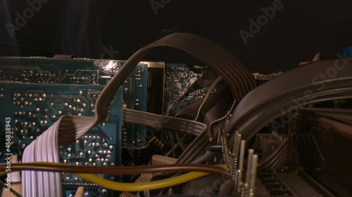 Sitting above a transformer with prongs and wires, wide angle macro of a number of printed circuit boards. Steady stream of smoke is rising near a back board.  Series of ribbon cables on the right. photo