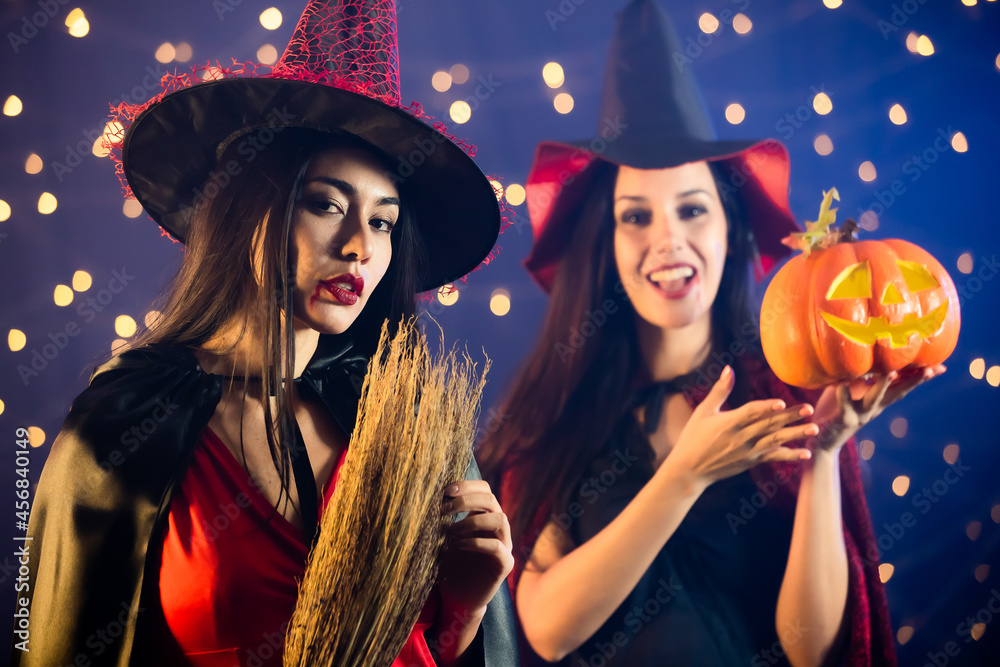 Portrait close up studio shot of Caucasian female in black red witch dress costume tall hat and shawl holding pumpkin face look at camera in front another Asian with broomstick in blurred background Portrait close up studio shot of Caucasian female in black red witch dress costume tall hat and shawl holding pumpkin face look at camera in front another Asian with broomstick in blurred background