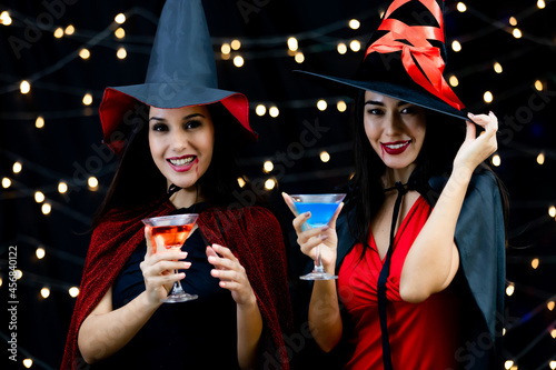 Caucasian and Asian sexy female in black and red witch dress costume tall hat and shawl holding glass of cocktail cheers and happy together on light bokeh background in Halloween party