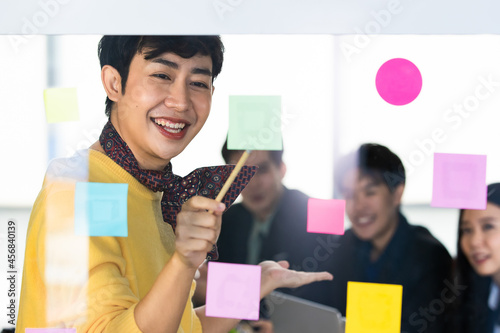 Asian trans woman on yellow shirt and scarf enjoy presenting business design to cheerful LGBT colleagues by point to colorful sticky paper note on glass board to explain creative strategy