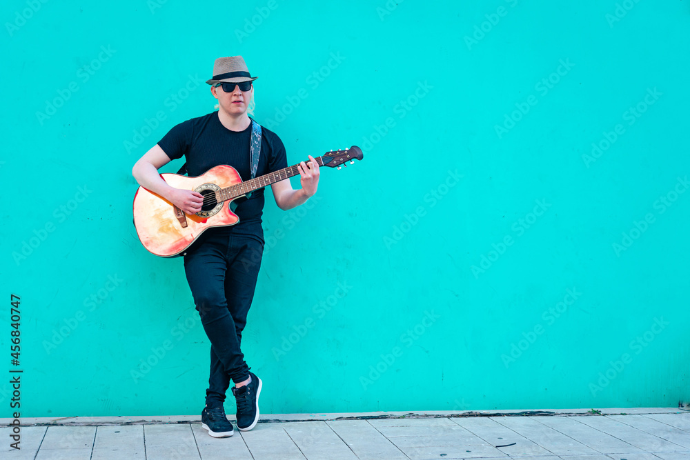 Obraz premium Close-up of albino man with hat playing guitar leaning against a colored wall. Horizontal orientation. Relaxation and inner peace concept.