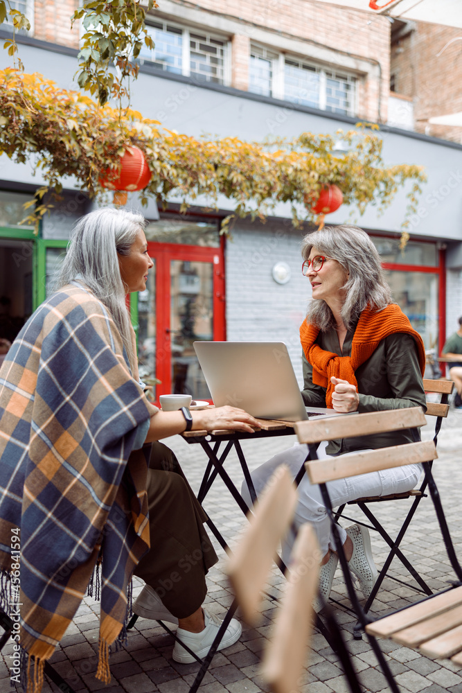 Mature women business partners with modern laptop discuss project at small table on outdoors cafe terrace on nice autumn day