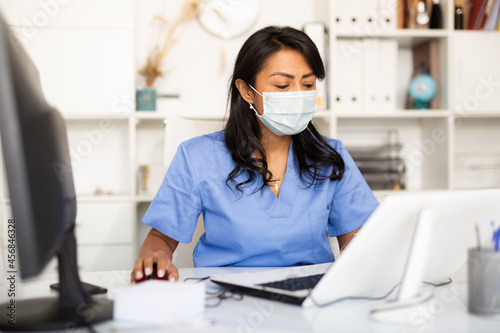Experienced female physician in protective mask filling up medical forms on laptop while sitting at table in office