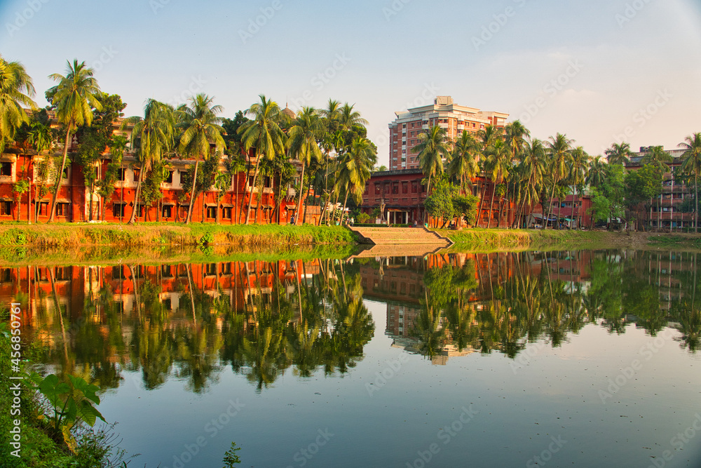 Obraz premium Coconut trees, red buildings, lake. Beautiful campus landscape. Reflection of the campus building on the lake. University of Dhaka, Bangladesh. November 2019