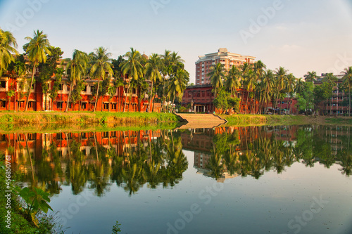 Coconut trees, red buildings, lake. Beautiful campus landscape. Reflection of the campus building on the lake. University of Dhaka, Bangladesh. November 2019