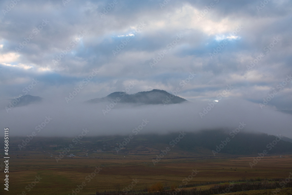 Obraz premium In the evening, the fog surrounds the trees, houses and mountains in the valley. Phobjikha Valley. One of the most lovely and high altitude river valleys, Bhutan. 2019