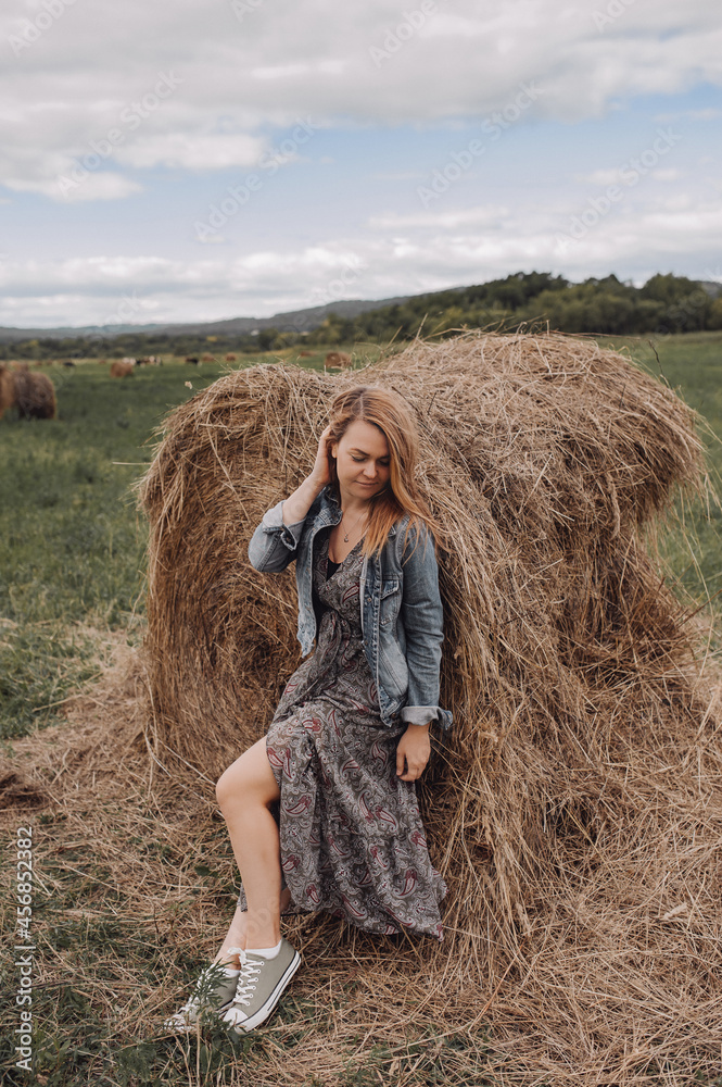 young beautiful woman in long dress, denim jacket, with bare legs in sneakers near large haystack in an empty field. local tourism.autumn nature landscape. enjoying nature. selective focus