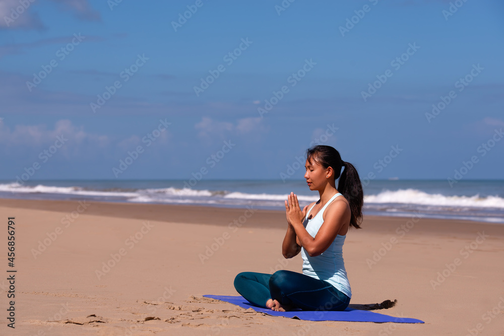 people, fitness, sport and healthy lifestyle concept - young asian woman making yoga pose hands together on tropical beach with blue sky background