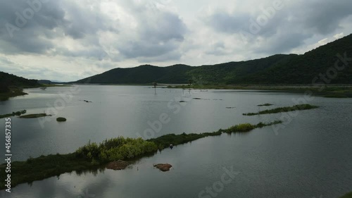 Aerial descending footage revealing this gorgeous lake with mountains around it, great sun setting with rainclouds, a man in the water setting up its fishing net; Saraburi, Thailand. photo