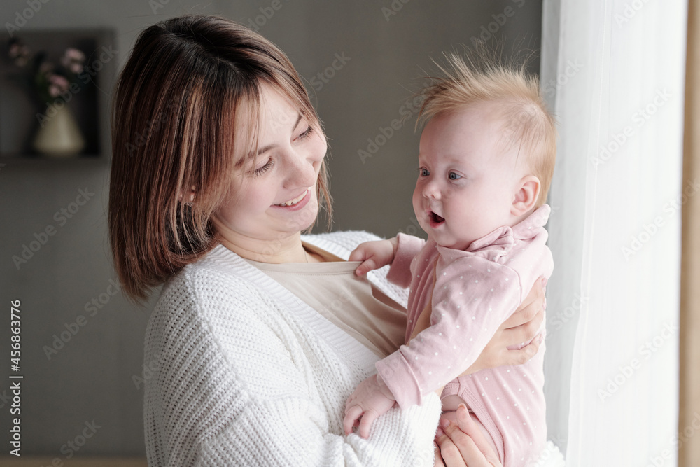 Young smiling female looking at adorable infant on her arms while standing in front of camera Young smiling female looking at adorable infant on her arms while standing in front of camera