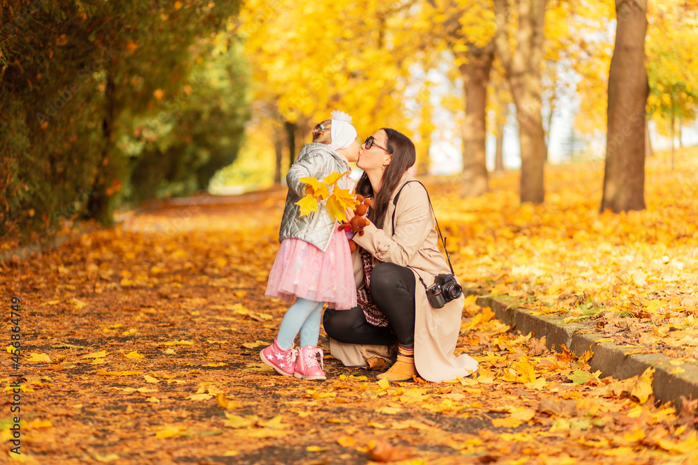 Fototapeta premium Mom and daughter in fashionable clothes walk in the park and make a kiss on the background of yellow autumn foliage