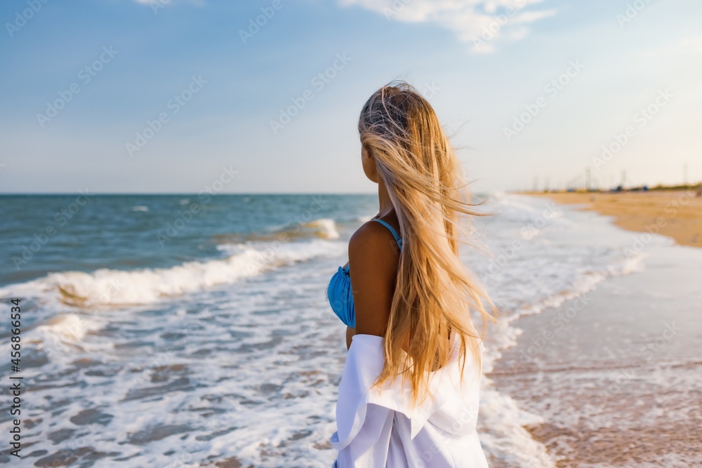 A slender girl in a gentle blue swimsuit and shirt walks along the sandy beach near the blue sea A slender girl in a gentle blue swimsuit and shirt walks along the sandy beach near the blue sea