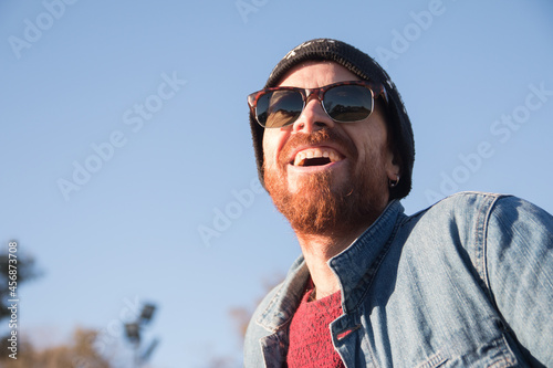 Closeup shot of a Caucasian handsome man with beard and sunglasses laughing under the sun