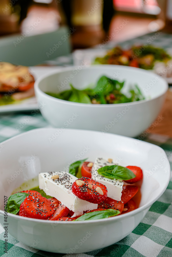 Caprese salad with strawberry, mozzarells, basil and balsamic sauce served in a white bowl over green plaid background. Caprese salad with strawberry, mozzarells, basil and balsamic sauce served in a white bowl over green plaid background.