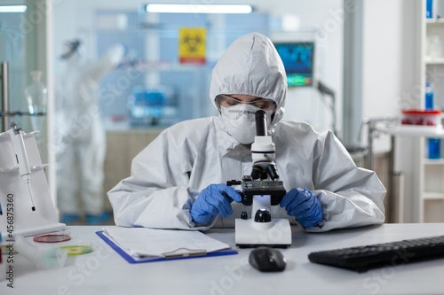 Biologist researcher wearing ppe medical suit analyzing genetics sample using clinical microscope in microbiology hospital laboratory. Scientist woman working at vaccine development expertise