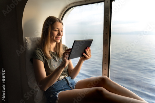 young woman seated in front of a porthole reading a digital book