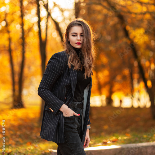 Young beautiful fashionable woman model with red lips in a trendy business suit with a fashion blazer, sweater and jeans walks in autumn park with colored fall orange foliage at sunset