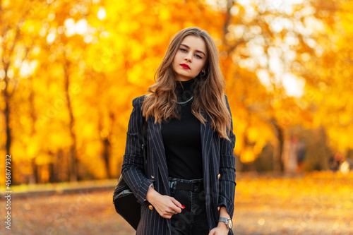 Autumn portrait of a pretty young woman with red lips in a black elegant suit with a blazer and sweater walks in the park with colored golden fall foliage at sunset