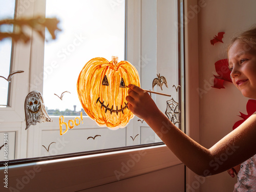 Child painting pumpkin on window preparing to celebrate Halloween. Little girl decorates her room autumn holiday at home