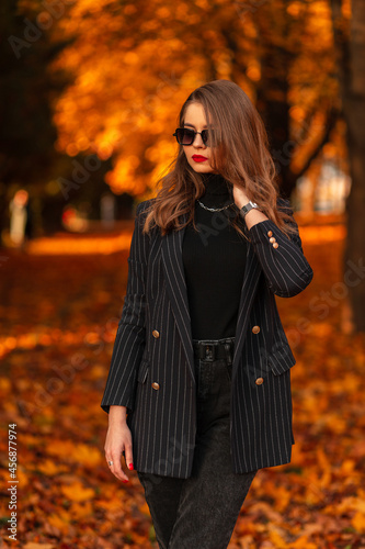 Beautiful young stylish business woman with cool sunglasses in a fashionable black suit with a elegant blazer and a sweater walks in an autumn park with colored golden foliage