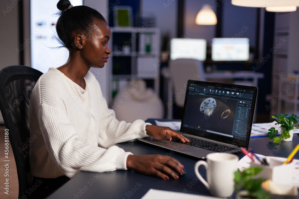 Focused african american engineer woman working at industrial gear prototype late at night in startup business company office. Engineering product construction idea on laptop screen Focused african american engineer woman working at industrial gear prototype late at night in startup business company office. Engineering product construction idea on laptop screen