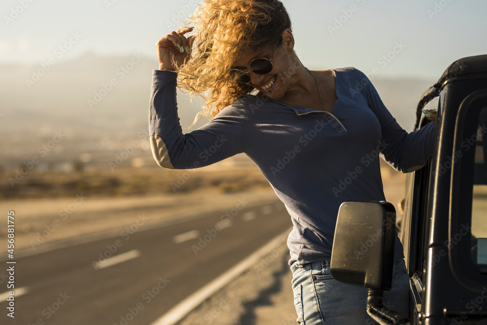 Fototapeta premium Cheerful woman in sunglasses hanging on side door of car at roadside. Young woman enjoying vacation on road trip. Carefree woman having fun leaning on outside of car door on her vacation trip
