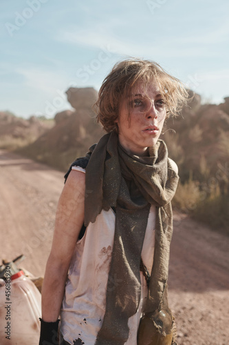 Young woman with dirty face and in torn clothes carrying cart behind her while walking in the desert