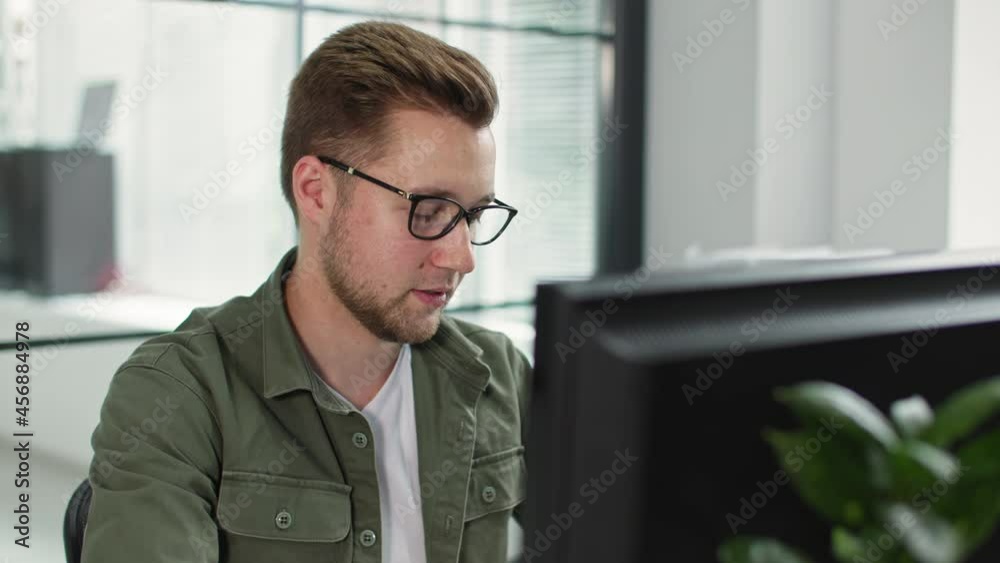 charming man in glasses for vision works at computer while sitting at table in office background of window charming man in glasses for vision works at computer while sitting at table in office background of window
