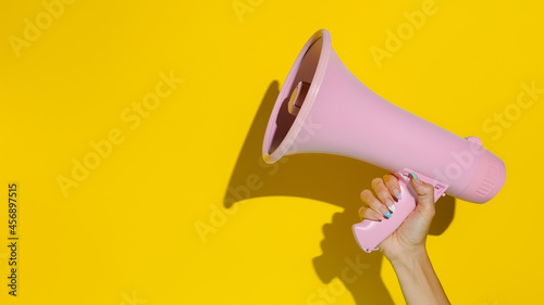 A woman's hand holding a pink megaphone isolated on a yellow background. Creative announcement concept. Loud voice of women. Women's rights and voice. Advertisement mock up with copy space for text. © Aleksandar