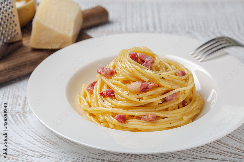 Spaghetti carbonara in a white plate. Pasta, pancetta and sauce made of egg yolk and parmesan cheese, black pepper. Hard cheese on background. Spaghetti carbonara in a white plate. Pasta, pancetta and sauce made of egg yolk and parmesan cheese, black pepper. Hard cheese on background.