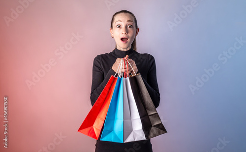 Woman with surprised face in a black dress is holding a colorful paper bags with purchases from a shop in her hands on a blue pink background. Concept of Black Friday, sale and shopping