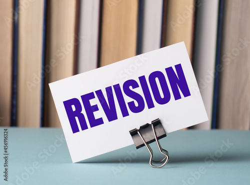 A white card with the text REVISION stands on a clip for papers on the table against the background of books. Defocus photo