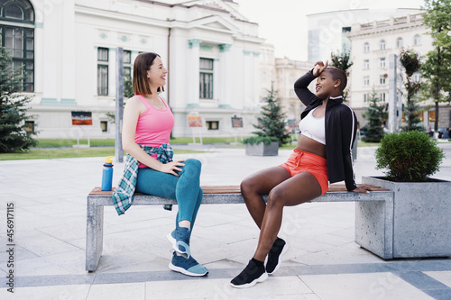 Cheerful smiling friends in sportswear sitting on bench in the city dicussing in park. Multiethnic women having a fitness workout break. photo