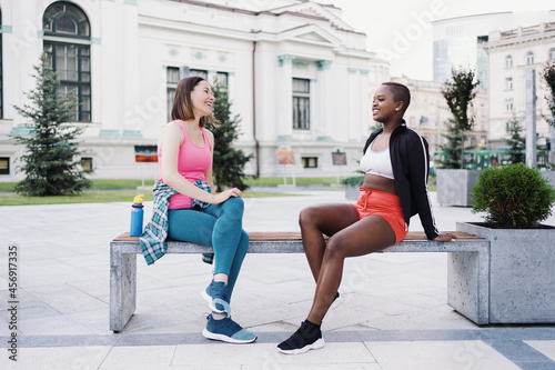 Cheerful smiling friends in sportswear sitting on bench in the city dicussing in park. Multiethnic women having a fitness workout break. photo