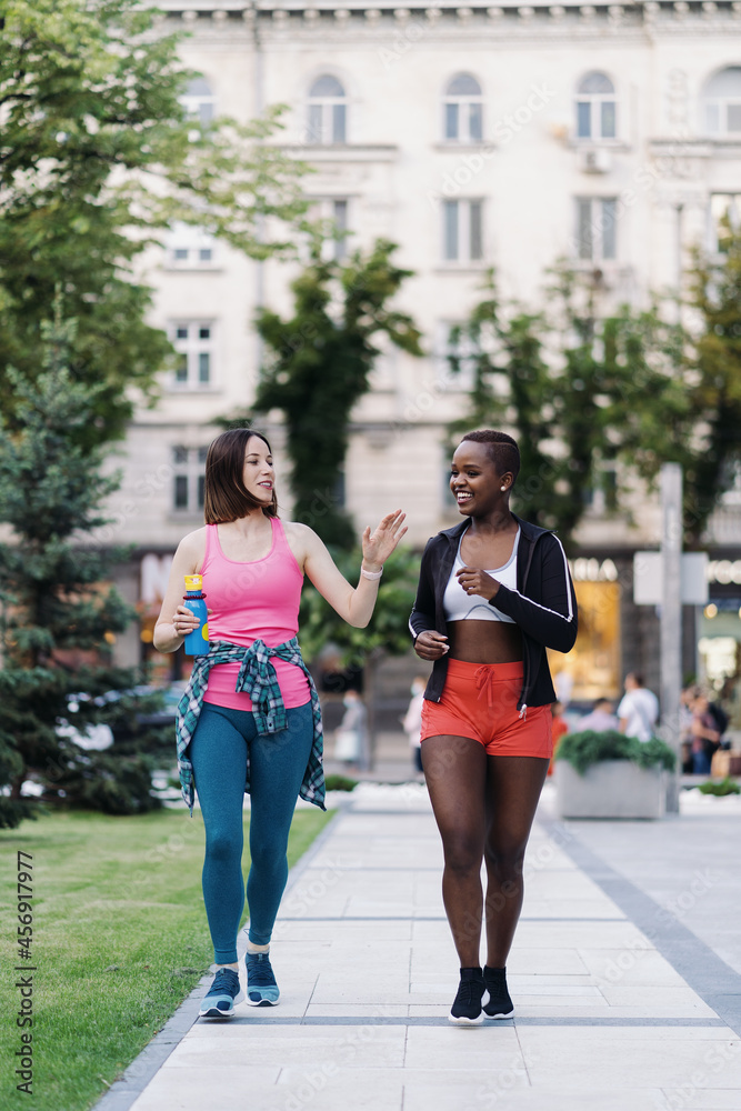 Cheerful smiling friends in sportswear walking after a sport session in the city discussing. Multiethnic women having a fitness workout jogging.