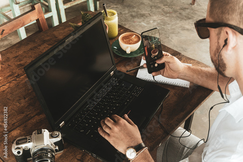 Człowiek pracujący przy laptopie z smartfonem, piszący notatki w zeszycie przy stole.