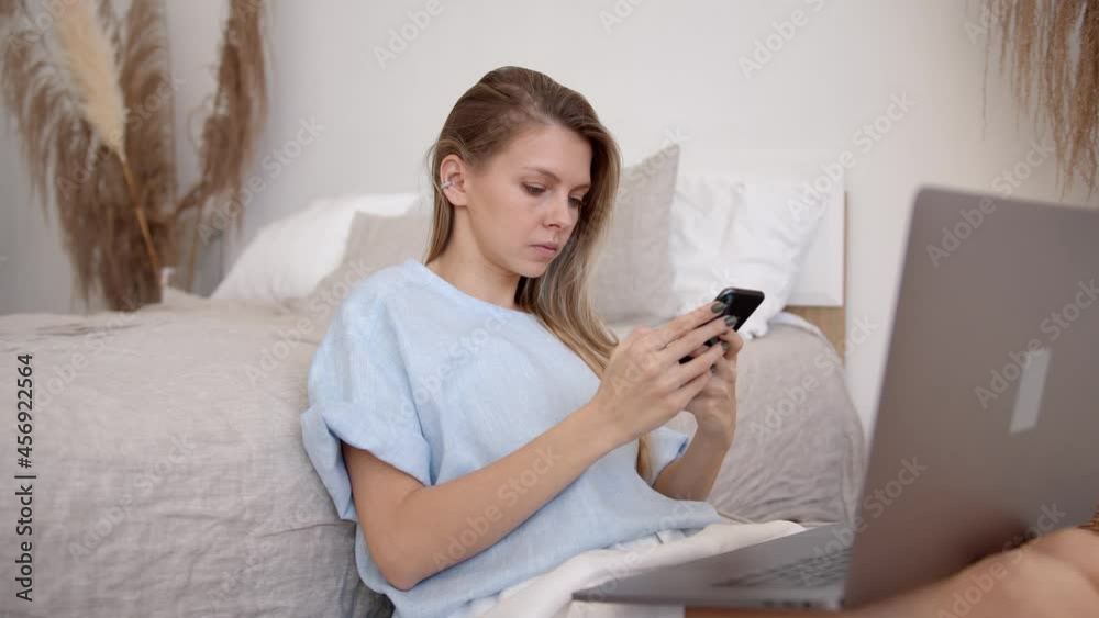 Blonde woman with cellphone in hands typing, sitting on the floor in bedroom. Side view of busy freelancer with devices working at home. Concept of remote work Blonde woman with cellphone in hands typing, sitting on the floor in bedroom. Side view of busy freelancer with devices working at home. Concept of remote work