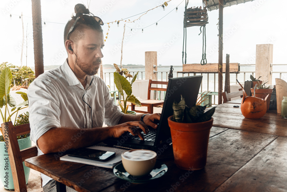 Mężczyzna, cyfrowy nomada pracujący z laptopem i telefonem przy stoliku w kawiarni, praca zdalna podczas podróży. - obrazy, fototapety, plakaty 