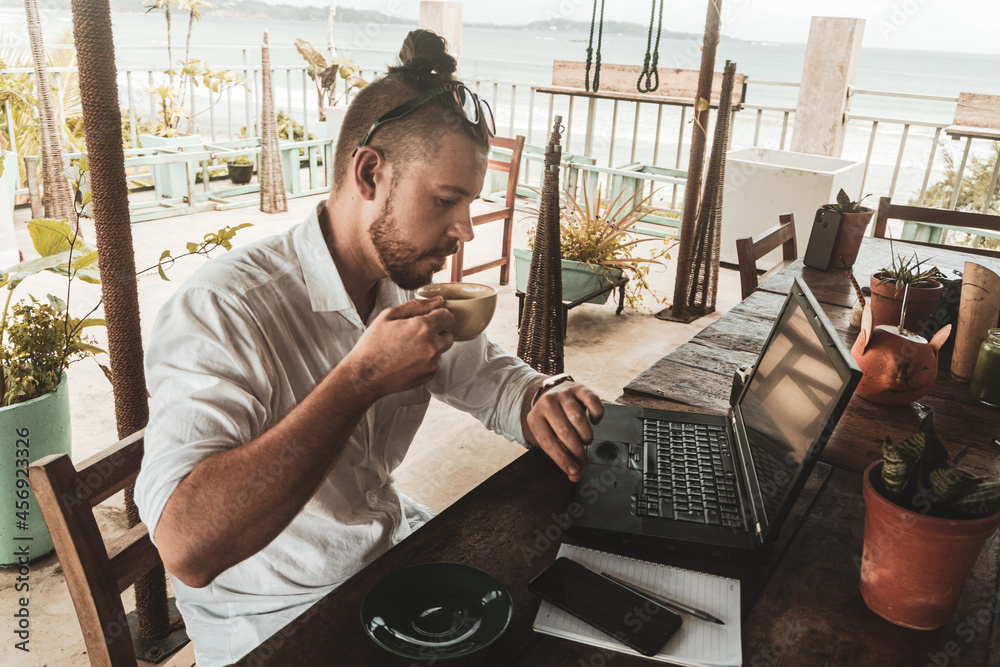 Mężczyzna, cyfrowy nomada pracujący z laptopem i telefonem przy stoliku w kawiarni, praca zdalna podczas podróży. - obrazy, fototapety, plakaty 