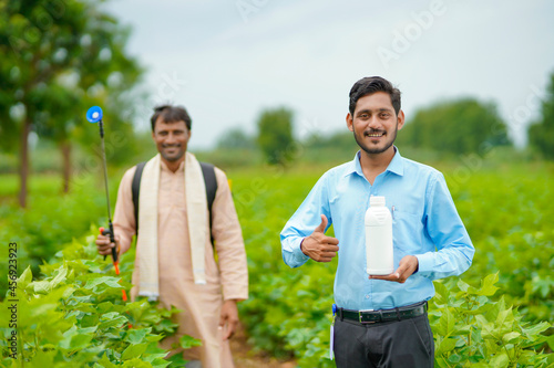 Young indian agronomist giving liquid fertilizer bottle to farmer and saying product information at green agriculture field.