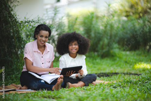 Cheerful young beautiful mother sitting with son with curly hair in park and helping him while using digital tablet while studying