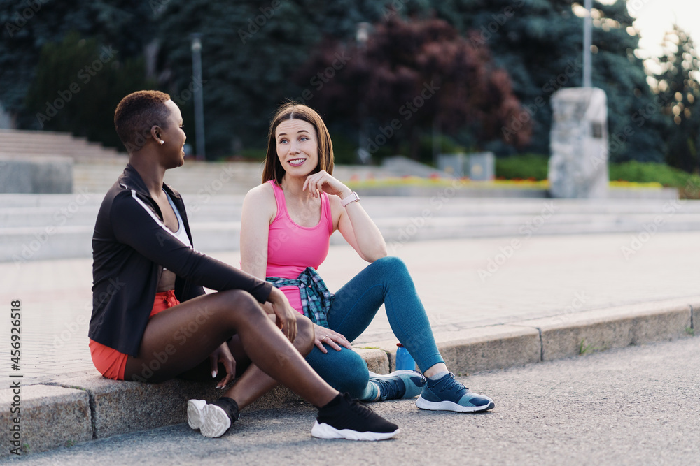 Cheerful smiling friends in sportswear sitting in the city dicussing. Multiethnic women having a fitness workout break.