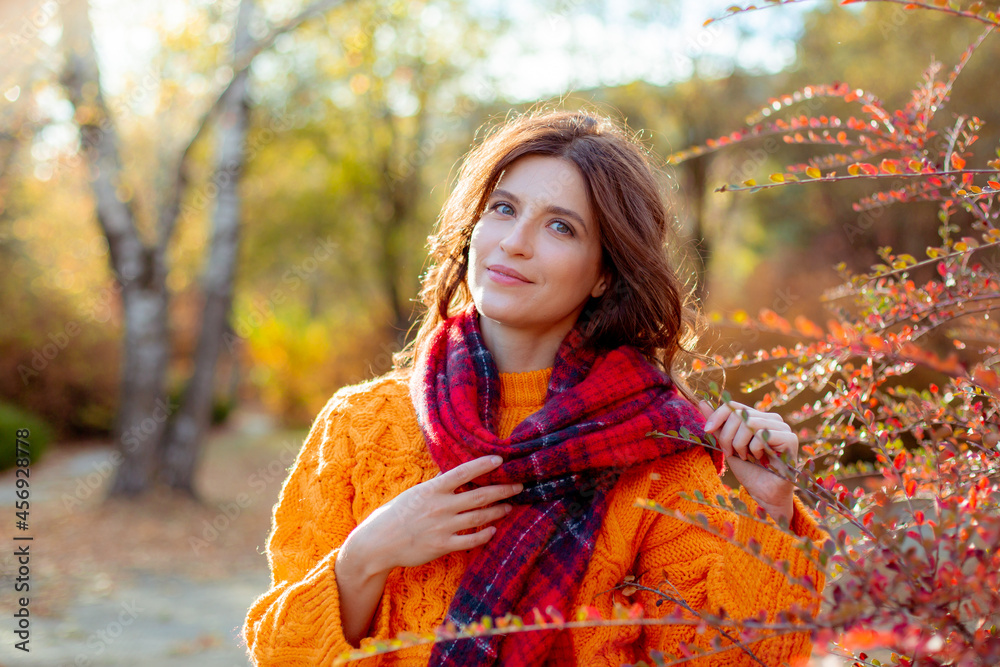 a young woman in an orange sweater wraps herself in a scarf in an autumn park