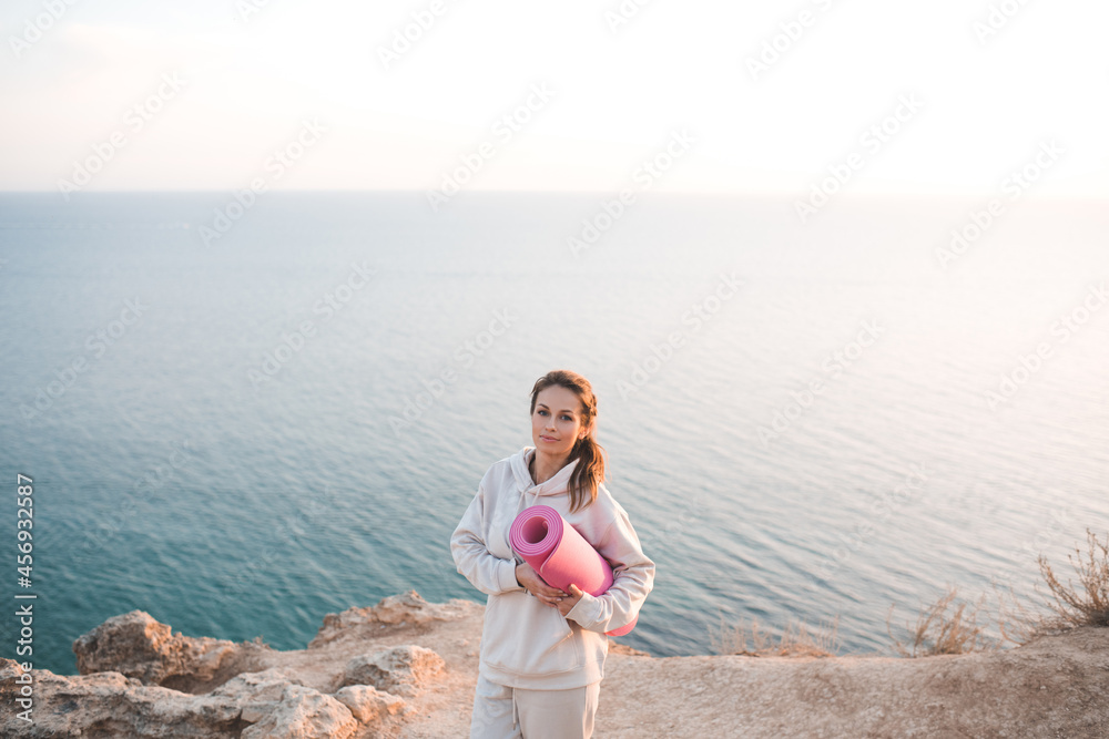 Smiling cheerful woman 25-26 year old hold yoga mat wear training hoodie posing over sea nature background. Healthy active lifestyle. Healthcare. Adult girl practice fitness outdoors. Happiness. 20s.