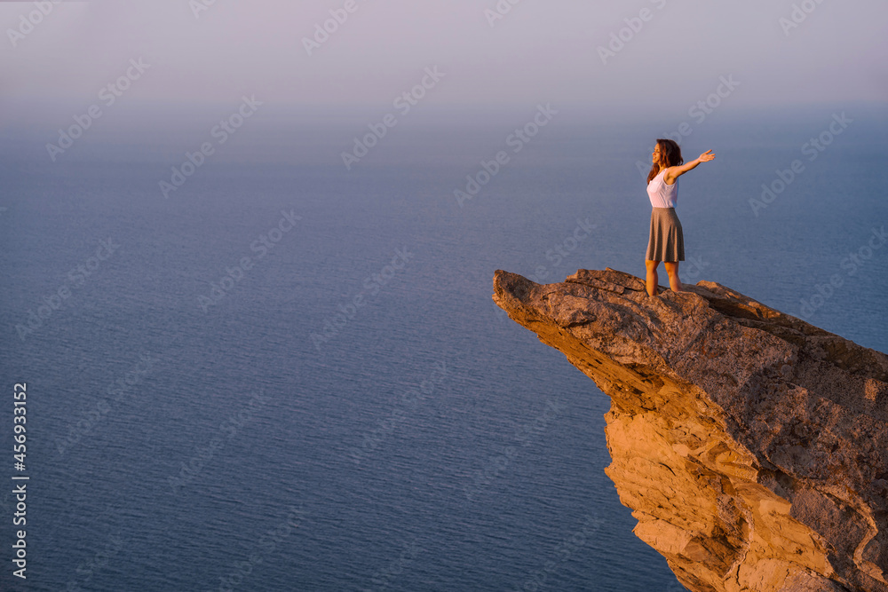 A young woman stands on a picturesque steep cliff above the sea against the sky. The concept of travel and freedom.