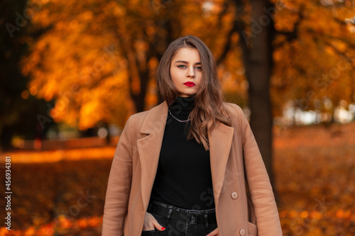 Pretty happy girl with red lips in a fashionable vintage beige coat with a sweater walks in an autumn park with orange leaves