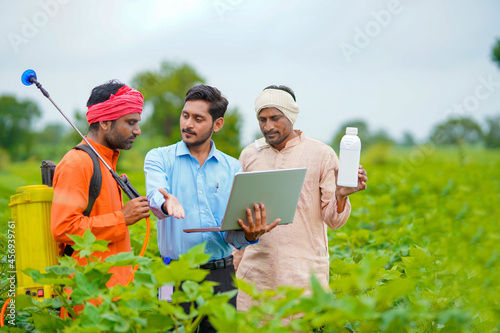 Young indian agronomist giving liquid fertilizer bottle to farmer and showing product information in laptop at green agriculture field.