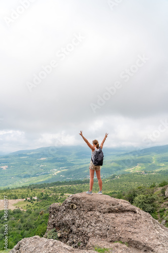 Happy woman with backpack standing on edge of cliff and looking at valley with her hands raised. Summer landscape with sporty girl, green grass, forest, hills, blue sky