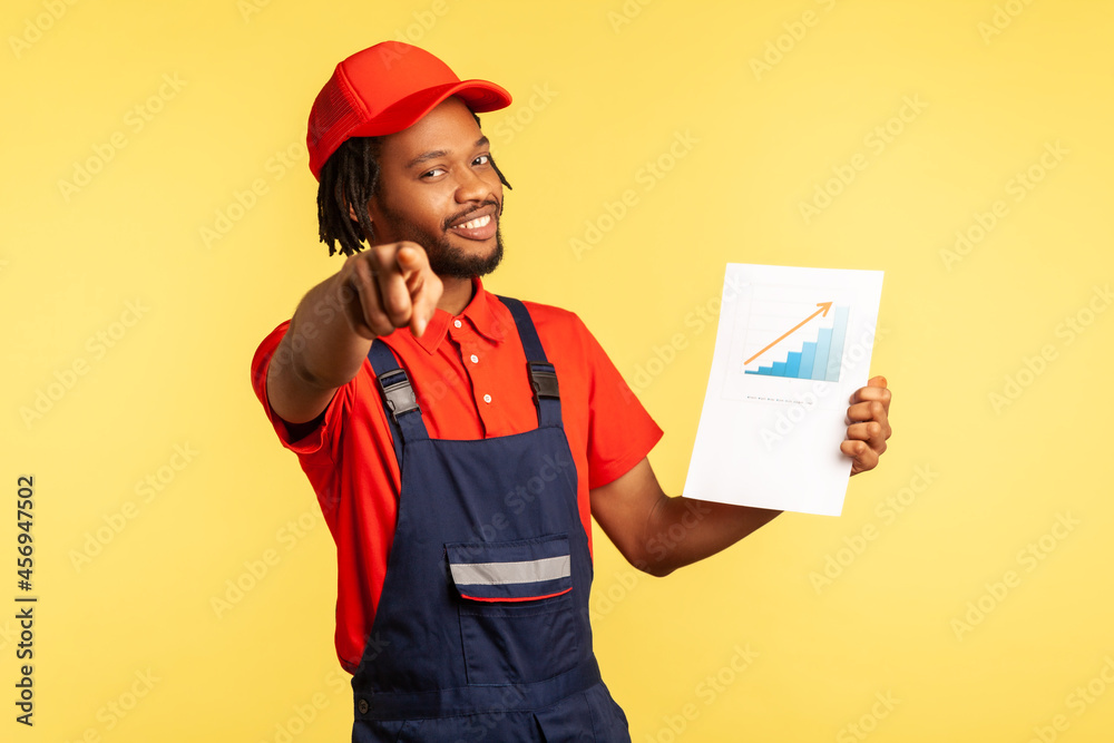Positive handyman in overalls holding paper chart showing increase of workers salaries, pointing to camera, inviting you to work in service industry. Indoor studio shot isolated on yellow background.