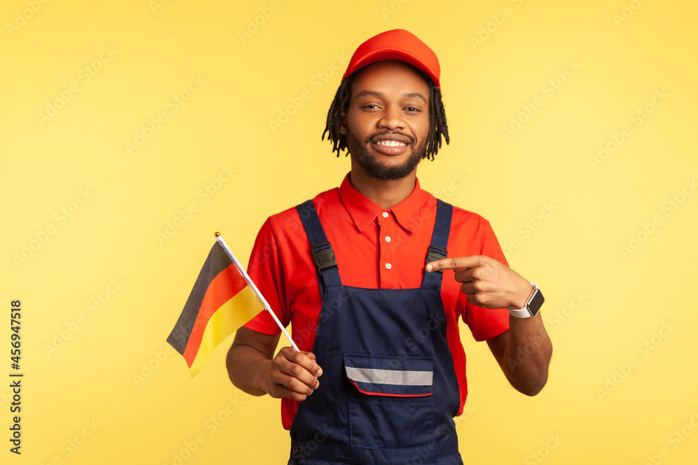 Satisfied handsome handyman in uniform with dreadlocks pointing finger at German flag in his hand and looking at camera with toothy smile. Indoor studio shot isolated on yellow background. Satisfied handsome handyman in uniform with dreadlocks pointing finger at German flag in his hand and looking at camera with toothy smile. Indoor studio shot isolated on yellow background.